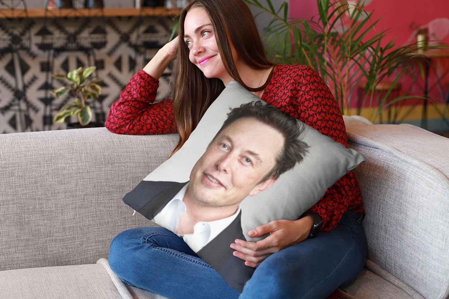 A young woman on a sofa holding an Elon Musk pillow and trying not to laugh.