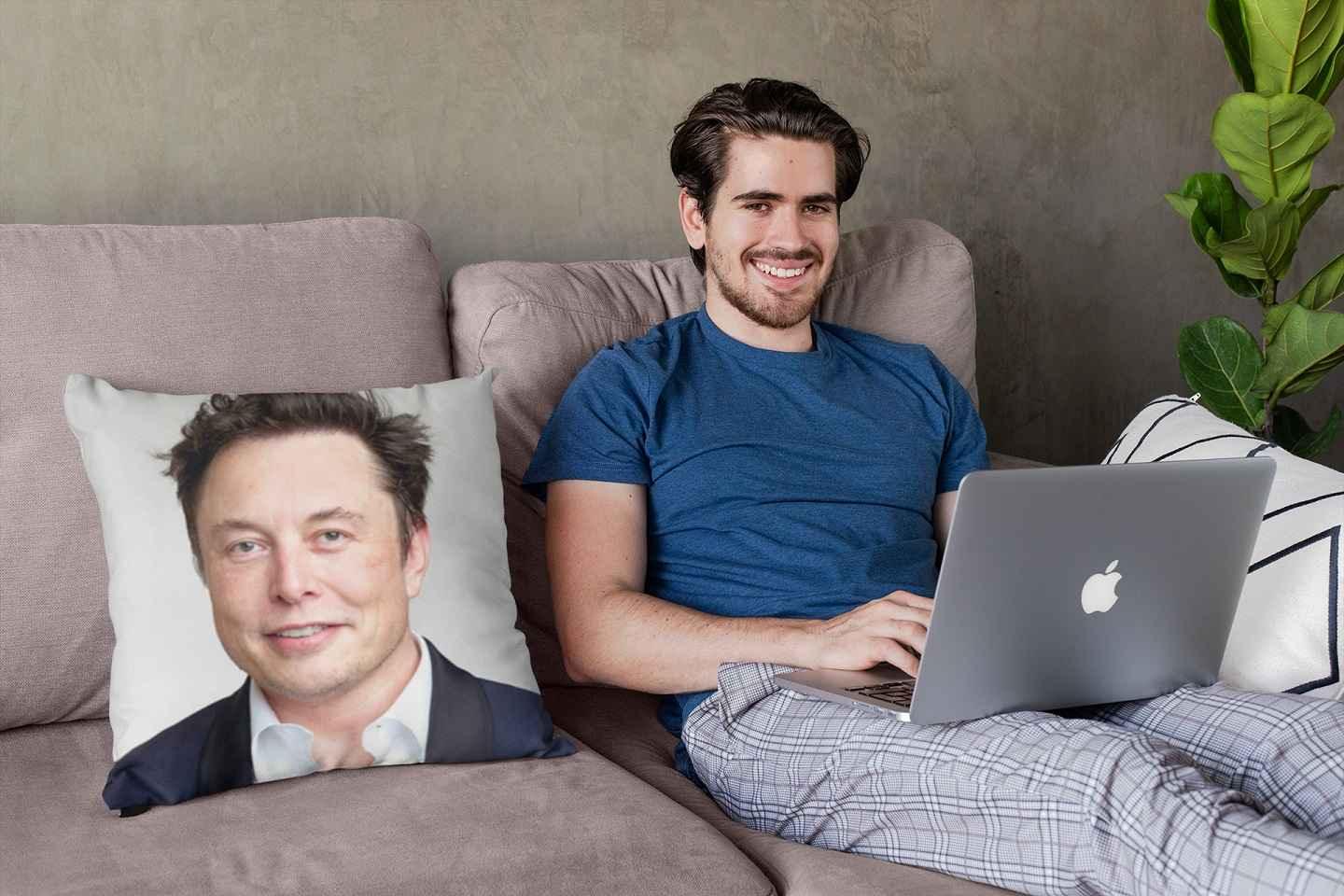 A smiling young man with a laptop sitting next to an Elon Musk pillow.