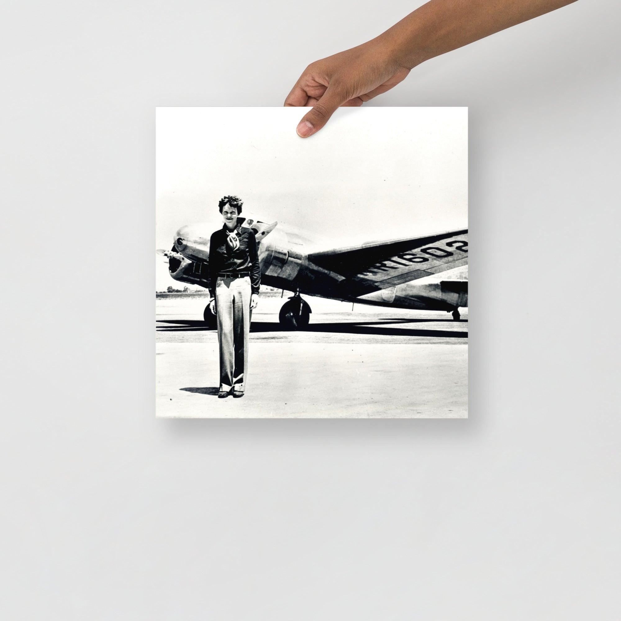 An Amelia Earhart standing in front of the Lockheed Electra on a plain backdrop in size 14x14”.